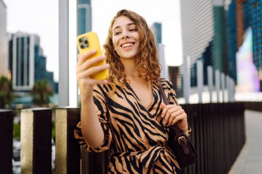 Elinde telefon olan genç bir kadın, şehir merkezine bakan yaya köprüsünde duruyor. Bir turist yürüyüş yapmaktan, mesajlaşmaktan ya da dışarıda blog yazmaktan hoşlanır. Günlük konsepti, yürüyüş.