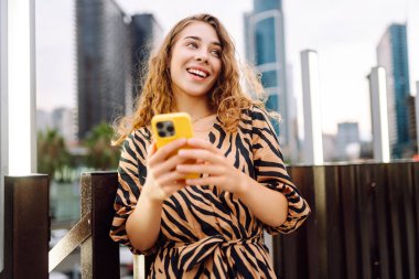 Elinde telefon olan genç bir kadın, şehir merkezine bakan yaya köprüsünde duruyor. Bir turist yürüyüş yapmaktan, mesajlaşmaktan ya da dışarıda blog yazmaktan hoşlanır. Günlük konsepti, yürüyüş.