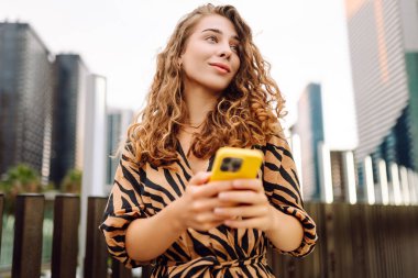 Elinde telefon olan genç bir kadın, şehir merkezine bakan yaya köprüsünde duruyor. Bir turist yürüyüş yapmaktan, mesajlaşmaktan ya da dışarıda blog yazmaktan hoşlanır. Günlük konsepti, yürüyüş.