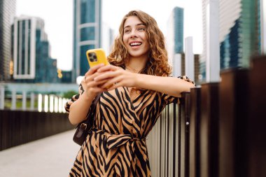 Elinde telefon olan genç bir kadın, şehir merkezine bakan yaya köprüsünde duruyor. Bir turist yürüyüş yapmaktan, mesajlaşmaktan ya da dışarıda blog yazmaktan hoşlanır. Günlük konsepti, yürüyüş.