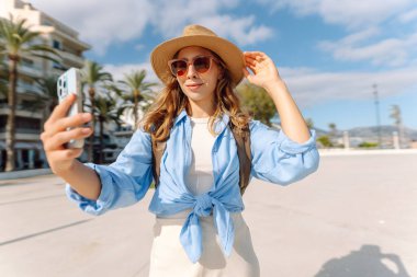 Şapkalı güzel bir turist elinde telefon ile şehir caddesinde duruyor. Genç bir kadın telefonuyla selfie çekiyor ve güneşli bir günde deniz manzarasından hoşlanıyor. Turizm, teknoloji ve kentselliğin kavramsal görüntüleri.