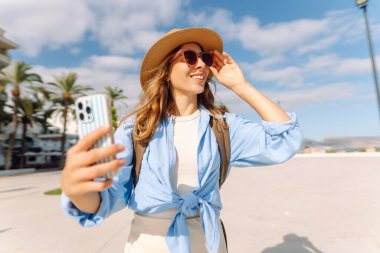 Şapkalı güzel bir turist elinde telefon ile şehir caddesinde duruyor. Genç bir kadın telefonuyla selfie çekiyor ve güneşli bir günde deniz manzarasından hoşlanıyor. Turizm, teknoloji ve kentselliğin kavramsal görüntüleri.