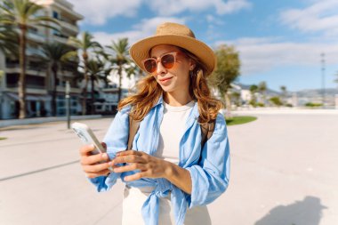 Şapkalı güzel bir turist elinde telefon ile şehir caddesinde duruyor. Genç bir kadın telefonuyla selfie çekiyor ve güneşli bir günde deniz manzarasından hoşlanıyor. Turizm, teknoloji ve kentselliğin kavramsal görüntüleri.