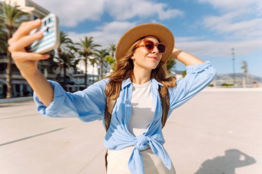 Şapkalı güzel bir turist elinde telefon ile şehir caddesinde duruyor. Genç bir kadın telefonuyla selfie çekiyor ve güneşli bir günde deniz manzarasından hoşlanıyor. Turizm, teknoloji ve kentselliğin kavramsal görüntüleri.