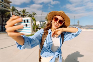 Şapkalı güzel bir turist elinde telefon ile şehir caddesinde duruyor. Genç bir kadın telefonuyla selfie çekiyor ve güneşli bir günde deniz manzarasından hoşlanıyor. Turizm, teknoloji ve kentselliğin kavramsal görüntüleri.