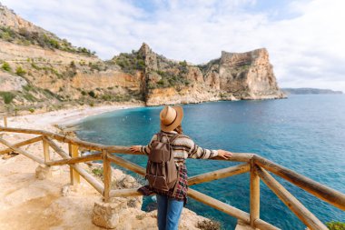 Şapka takan ve sırt çantası taşıyan genç bir kadın yürüyüşçü, denize ve kayalıklara bakan güzel bir bakış açısında duruyor. Neşeli bir kadın yürüyüş yapıyor, özgür hissediyor ve güneşli günün tadını çıkarıyor..
