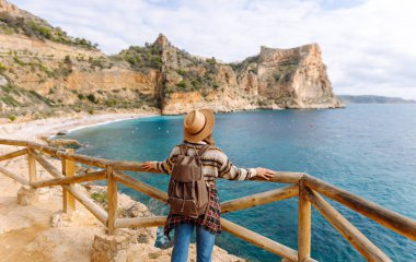 Şapka takan ve sırt çantası taşıyan genç bir kadın yürüyüşçü, denize ve kayalıklara bakan güzel bir bakış açısında duruyor. Neşeli bir kadın yürüyüş yapıyor, özgür hissediyor ve güneşli günün tadını çıkarıyor..