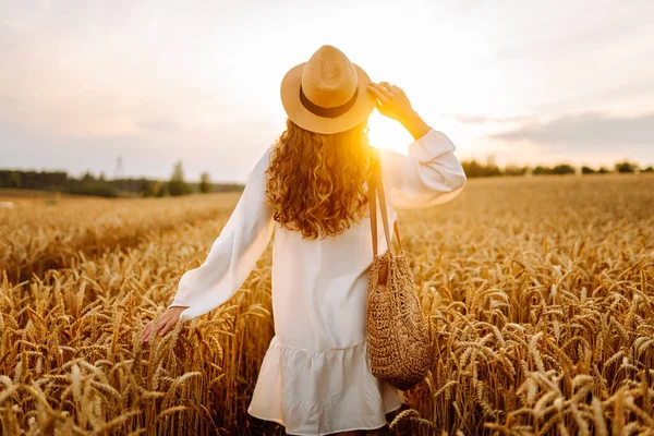 A sweet woman in a hat stands in a wheat field at sunset. A beautiful woman enjoys the sunset and strolls through a farmland. Concept of beauty, nature.