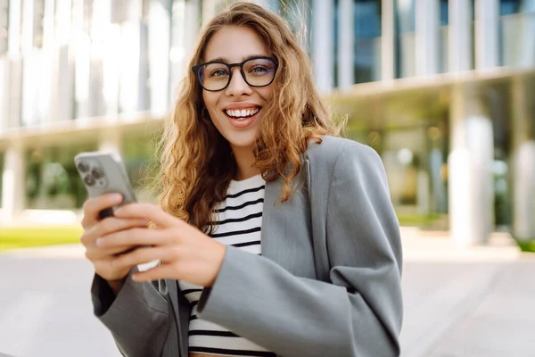 Gözlük takan neşeli bir kadının portresi ve modern bir ofis binasının arka planında telefon kullanmak. Şık bir kadın blogu yapıyor ve güneşli bir gün geçiriyor..