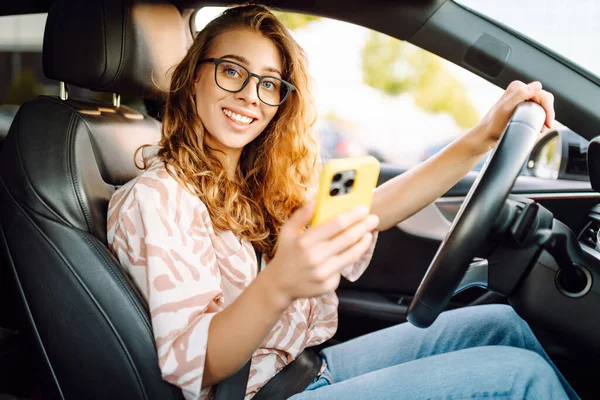 Araba kullanırken telefonla konuşan gözlüklü genç bir kadının portresi. Güneşli bir günde modern bir arabada elinde telefon tutan bir kadın sürücü. Ulaşım ve teknoloji kavramı.