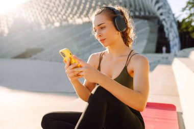 Atletik bir kadın parkta yoga minderinde oturuyor, kablosuz kulaklık takıyor ve telefonunu kontrol ediyor. Spor kıyafetleri içindeki genç bir kadın dışarıda antrenman yapmadan önce ısınıyor. Konsept: spor, müzik.