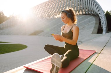Atletik bir kadın parkta yoga minderinde oturuyor, kablosuz kulaklık takıyor ve telefonunu kontrol ediyor. Spor kıyafetleri içindeki genç bir kadın dışarıda antrenman yapmadan önce ısınıyor. Konsept: spor, müzik.