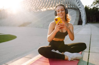 Atletik bir kadın parkta yoga minderinde oturuyor, kablosuz kulaklık takıyor ve telefonunu kontrol ediyor. Spor kıyafetleri içindeki genç bir kadın dışarıda antrenman yapmadan önce ısınıyor. Konsept: spor, müzik.