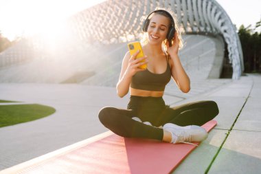 Atletik bir kadın parkta yoga minderinde oturuyor, kablosuz kulaklık takıyor ve telefonunu kontrol ediyor. Spor kıyafetleri içindeki genç bir kadın dışarıda antrenman yapmadan önce ısınıyor. Konsept: spor, müzik.