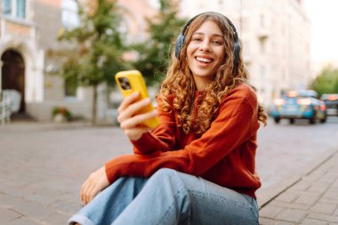 Kablosuz kulaklıkları ve telefonu olan neşeli bir kadın güneşli bir günde şehir ortamının tadını çıkarıyor. Genç bir kadın müzik dinler ve eski kasabada gezinir. Dinlenme ve rahatlama kavramı.