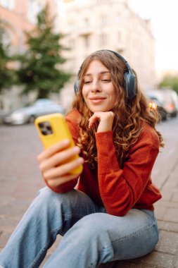 Kablosuz kulaklıkları ve telefonu olan neşeli bir kadın güneşli bir günde şehir ortamının tadını çıkarıyor. Genç bir kadın müzik dinler ve eski kasabada gezinir. Dinlenme ve rahatlama kavramı.