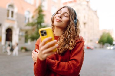 Kablosuz kulaklıkları ve telefonu olan neşeli bir kadın güneşli bir günde şehir ortamının tadını çıkarıyor. Genç bir kadın müzik dinler ve eski kasabada gezinir. Dinlenme ve rahatlama kavramı.