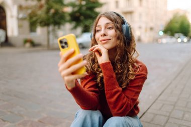 Kablosuz kulaklıkları ve telefonu olan neşeli bir kadın güneşli bir günde şehir ortamının tadını çıkarıyor. Genç bir kadın müzik dinler ve eski kasabada gezinir. Dinlenme ve rahatlama kavramı.