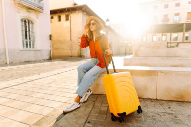 Güneşli bir günde, sarı bavulu ve sırt çantasıyla şehir caddesinde yürüyen güzel bir gezgin. Güneş gözlüklü ve şapkalı genç bayan gezintiden hoşlanıyor. Turizm ve eğlence kavramı.