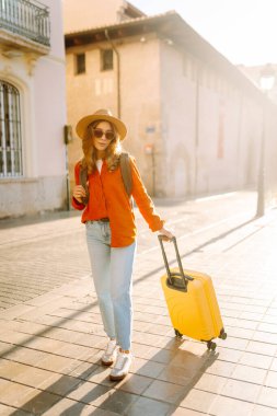 Güneşli bir günde, sarı bavulu ve sırt çantasıyla şehir caddesinde yürüyen güzel bir gezgin. Güneş gözlüklü ve şapkalı genç bayan gezintiden hoşlanıyor. Turizm ve eğlence kavramı.