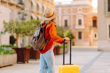 Güneşli bir günde, sarı bavulu ve sırt çantasıyla şehir caddesinde yürüyen güzel bir gezgin. Güneş gözlüklü ve şapkalı genç bayan gezintiden hoşlanıyor. Turizm ve eğlence kavramı.