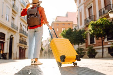 Güneşli bir günde, sarı bavulu ve sırt çantasıyla şehir caddesinde yürüyen güzel bir gezgin. Güneş gözlüklü ve şapkalı genç bayan gezintiden hoşlanıyor. Turizm ve eğlence kavramı.