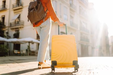 Güneşli bir günde, sarı bavulu ve sırt çantasıyla şehir caddesinde yürüyen güzel bir gezgin. Güneş gözlüklü ve şapkalı genç bayan gezintiden hoşlanıyor. Turizm ve eğlence kavramı.