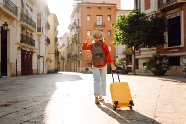 Güneşli bir günde, sarı bavulu ve sırt çantasıyla şehir caddesinde yürüyen güzel bir gezgin. Güneş gözlüklü ve şapkalı genç bayan gezintiden hoşlanıyor. Turizm ve eğlence kavramı.
