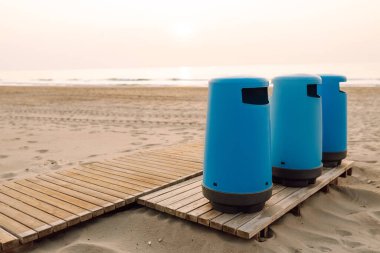 Üç mavi çöp tenekesi kumsaldaki ahşap bir platformda duruyor. Gün batımında halka açık bir sahilde çöp konteynırları. Konsept ekoloji, temizlik ve doğa..