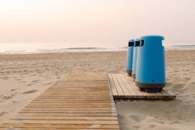 Üç mavi çöp tenekesi kumsaldaki ahşap bir platformda duruyor. Gün batımında halka açık bir sahilde çöp konteynırları. Konsept ekoloji, temizlik ve doğa..