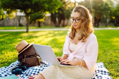 Genç bir kadın güneşli bir parkta dizüstü bilgisayarla battaniyenin üzerinde oturuyor. Dişi bir serbest çalışan dışarıda dizüstü bilgisayarda çalışıyor, doğanın tadını çıkarıyor. Uzaktan çalışma, rahatlama ve doğa kavramları.