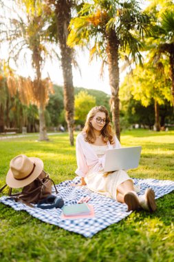 Genç bir kadın güneşli bir parkta dizüstü bilgisayarla battaniyenin üzerinde oturuyor. Dişi bir serbest çalışan dışarıda dizüstü bilgisayarda çalışıyor, doğanın tadını çıkarıyor. Uzaktan çalışma, rahatlama ve doğa kavramları.