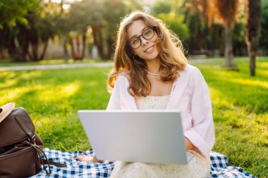 Genç bir kadın güneşli bir parkta dizüstü bilgisayarla battaniyenin üzerinde oturuyor. Dişi bir serbest çalışan dışarıda dizüstü bilgisayarda çalışıyor, doğanın tadını çıkarıyor. Uzaktan çalışma, rahatlama ve doğa kavramları.