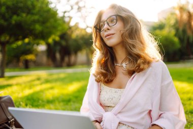 Genç bir kadın güneşli bir parkta dizüstü bilgisayarla battaniyenin üzerinde oturuyor. Dişi bir serbest çalışan dışarıda dizüstü bilgisayarda çalışıyor, doğanın tadını çıkarıyor. Uzaktan çalışma, rahatlama ve doğa kavramları.