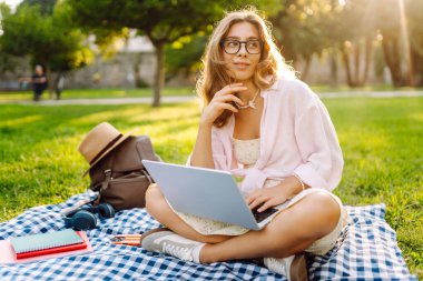Genç bir kadın güneşli bir parkta dizüstü bilgisayarla battaniyenin üzerinde oturuyor. Dişi bir serbest çalışan dışarıda dizüstü bilgisayarda çalışıyor, doğanın tadını çıkarıyor. Uzaktan çalışma, rahatlama ve doğa kavramları.