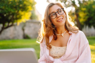 Genç bir kadın güneşli bir parkta dizüstü bilgisayarla battaniyenin üzerinde oturuyor. Dişi bir serbest çalışan dışarıda dizüstü bilgisayarda çalışıyor, doğanın tadını çıkarıyor. Uzaktan çalışma, rahatlama ve doğa kavramları.