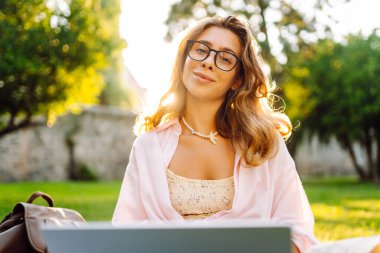 Genç bir kadın güneşli bir parkta dizüstü bilgisayarla battaniyenin üzerinde oturuyor. Dişi bir serbest çalışan dışarıda dizüstü bilgisayarda çalışıyor, doğanın tadını çıkarıyor. Uzaktan çalışma, rahatlama ve doğa kavramları.