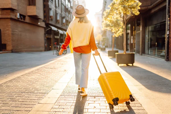 Güneşli bir günde, sarı bavulu ve sırt çantasıyla şehir caddesinde yürüyen güzel bir gezgin. Güneş gözlüklü ve şapkalı genç bayan gezintiden hoşlanıyor. Turizm ve eğlence kavramı.
