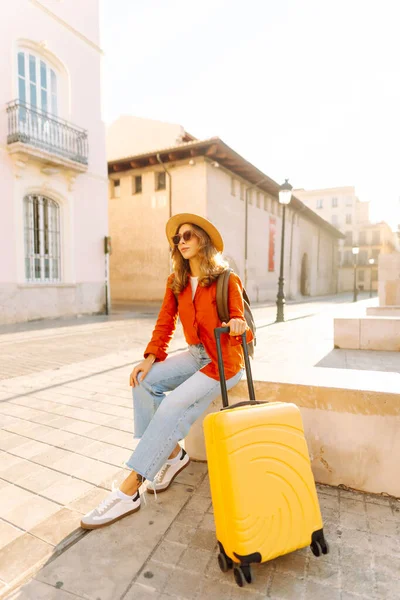 Güneşli bir günde, sarı bavulu ve sırt çantasıyla şehir caddesinde yürüyen güzel bir gezgin. Güneş gözlüklü ve şapkalı genç bayan gezintiden hoşlanıyor. Turizm ve eğlence kavramı.