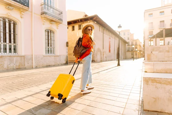 Güneşli bir günde, sarı bavulu ve sırt çantasıyla şehir caddesinde yürüyen güzel bir gezgin. Güneş gözlüklü ve şapkalı genç bayan gezintiden hoşlanıyor. Turizm ve eğlence kavramı.