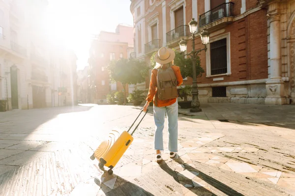 Güneşli bir günde, sarı bavulu ve sırt çantasıyla şehir caddesinde yürüyen güzel bir gezgin. Güneş gözlüklü ve şapkalı genç bayan gezintiden hoşlanıyor. Turizm ve eğlence kavramı.