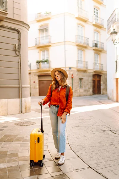 Güneşli bir günde, sarı bavulu ve sırt çantasıyla şehir caddesinde yürüyen güzel bir gezgin. Güneş gözlüklü ve şapkalı genç bayan gezintiden hoşlanıyor. Turizm ve eğlence kavramı.