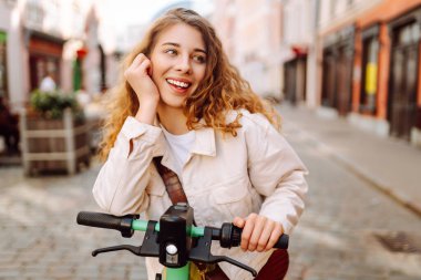 Neşeli bir kadın güneşli şehir caddelerinde elektrikli scooter kullanıyor. Genç bir turist modern bir scooterla gezmekten hoşlanıyor. Ekoloji ve yürüme kavramı.
