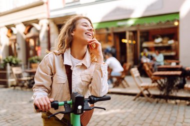 Neşeli bir kadın güneşli şehir caddelerinde elektrikli scooter kullanıyor. Genç bir turist modern bir scooterla gezmekten hoşlanıyor. Ekoloji ve yürüme kavramı.