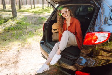 Güneşli bir ormanda sırt çantasıyla bir arabanın bagajında oturan güzel bir kadın. Güneş gözlüklü genç bir gezgin gün batımında doğanın tadını çıkarır. Kavram: ulaşım, macera.