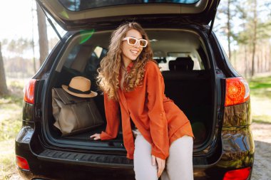 Güneşli bir ormanda sırt çantasıyla bir arabanın bagajında oturan güzel bir kadın. Güneş gözlüklü genç bir gezgin gün batımında doğanın tadını çıkarır. Kavram: ulaşım, macera.