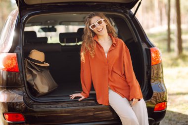 Güneşli bir ormanda sırt çantasıyla bir arabanın bagajında oturan güzel bir kadın. Güneş gözlüklü genç bir gezgin gün batımında doğanın tadını çıkarır. Kavram: ulaşım, macera.