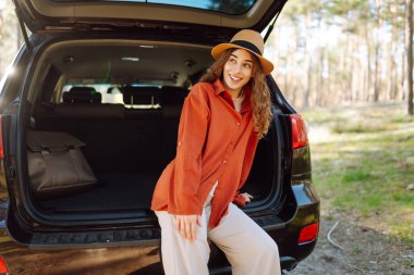 Güneşli bir ormanda sırt çantasıyla bir arabanın bagajında oturan güzel bir kadın. Güneş gözlüklü genç bir gezgin gün batımında doğanın tadını çıkarır. Kavram: ulaşım, macera.