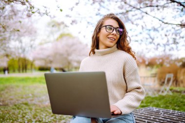 Gülen bir kadın lanet bir parkta dizüstü bilgisayarla masada oturuyor. Gözlük takan genç bir kadın gün batımında dışarıda dizüstü bilgisayarda çalışıyor. Uzaktan çalışma, rahatlama ve hafta sonları için kavramlar.