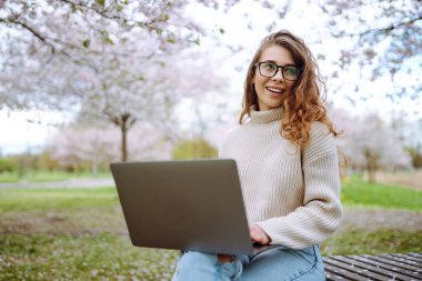 Gülen bir kadın lanet bir parkta dizüstü bilgisayarla masada oturuyor. Gözlük takan genç bir kadın gün batımında dışarıda dizüstü bilgisayarda çalışıyor. Uzaktan çalışma, rahatlama ve hafta sonları için kavramlar.